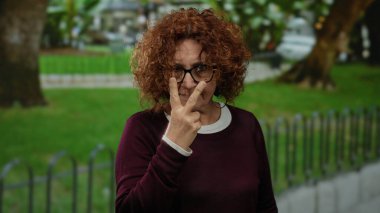 Parkta yoğun bir ifadeyle jestler yapan, kıvırcık saçlı, gözlüklü ve açık havada dikkat çeken bir kadın..