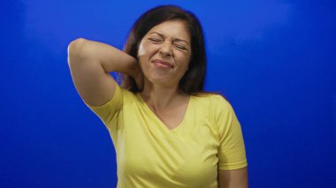 Sarı gömlekli bir kadın mavi stüdyoda gerginlik gösteren gözleri kapalı boynuna dokunuyor..