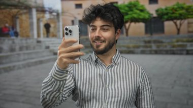 Genç adam, şık mimari ve yeşilliklerle çevrili, gösterişli İspanyol kültürü ve yaşam tarzını gösteren bir sokak ortamında akıllı telefonuyla selfie çekiyor..