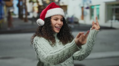 Noel Baba şapkası takan kadın, tatil sezonunda neşeli sokak ortamında neşeli jestler yapar. Neşeli giysiler ve açık havada hareketli davranışlar sergiler..