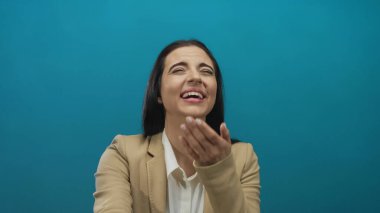 Bej takım elbiseli genç İspanyol kadın, gülüyor ve mavi bir duvar arkasına işaret ediyor, neşeli duyguları güvenli bir şekilde ifade ediyor..