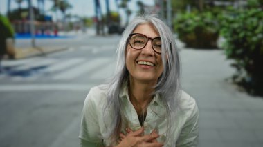 Yaşlı, kır saçlı, dövmeli bir kadın güneşli bir caddede sıcacık gülümsüyor, kaldırımda ağaçların oluşturduğu bir şehir ortamında neşe ve pozitiflik sergiliyor..