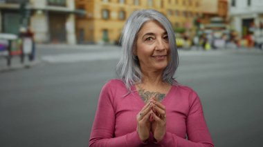 Pembe kazaklı yaşlı kadın hareketli bir sokakta neşeli jestler yapıyor. Arka planda bulanık binalar ve oradan geçen bir bisikletliyle canlı bir şehir manzarası yaratıyor..