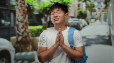 Sırt çantalı genç bir adam güneşli bir şehir caddesinde durup dua ederken palmiye ağaçları ve park halindeki arabaların arasında duygularını ifade ediyor..