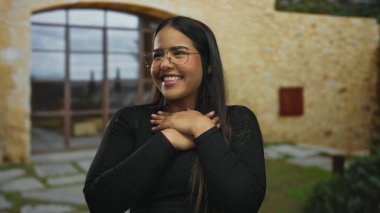 Güneşli bir günde kır evinin önünde kolları açık gülümseyen genç bir kadın. Gözlüklü ve siyah gömlekli. Mutluluğu iletiyor.