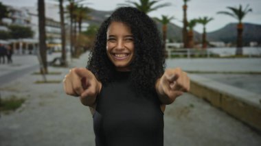 Siyah kıvırcık saçlı genç İspanyol kadın geniş bir gülümseme ve canlı caddede kamerayı işaret ediyor. Eğlenceli bir davet..