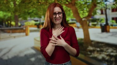 Kızıl saçlı kadın güneşli bir parkta duruyor, gözlük takıyor, elleriyle duygularını ifade ediyor, kırmızı bir bluz giyiyor, yeşil ağaçlar ve açık hava manzarası ile çevrili..