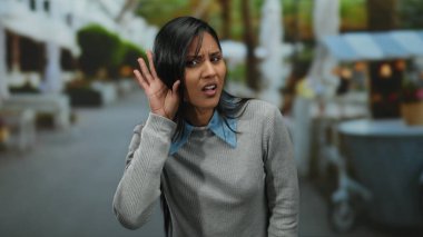 Sokakta jest dinleyen bir kadın. Latin kökenli, genç, gündüz giyimli, sıradan mahalle ağaçları.