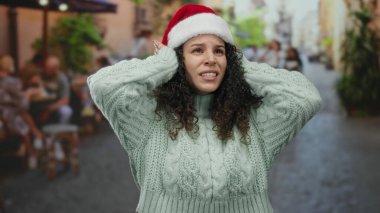 Noel Baba şapkası takan bir kadın, sokak arkası hareketli kafeterya terasında stres altında. Bu da tatil bunalımını ve şehir yaşamının zıtlığını akla getiriyor..