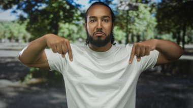 Beyaz tişörtlü sakallı adam orman düzeninde her iki işaret parmağı aşağı doğru, doğrudan kameraya bakın;.