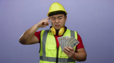 Miğfer takan genç bir adam mor bir arka plana karşı düşünceli bir ifadeyle bize dolar tutuyor..