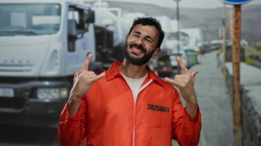 Turuncu hapishane tulumlu İspanyol bir adam inşaat alanında taş jestler yapıyor, arkada kamyonlarla neşeyle gülümsüyor, kaygısız bir mahkumu somutlaştırıyor..