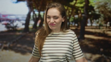 Çizgili gömlekli kadın, parlak güneş ışığı altında meşe ağaçlarının yanındaki yeşil parkta el ve kırışıklıklar silkeliyor..