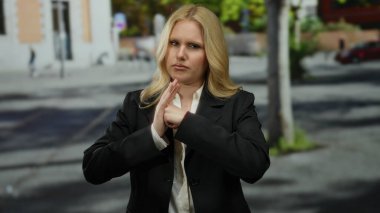 Şehirdeki genç sarışın kadın, şehir mimarisi ve arka planda bulanık manzarayla çevrili ciddi bir ifadeyle kavga jesti yapıyor..
