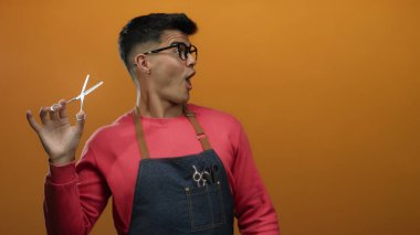 Gözlüklü genç İspanyol adam elinde makas tutarak canlı turuncu arka planda, saç şekillendirmedeki eğlence ve yaratıcılığı ifade ediyor..