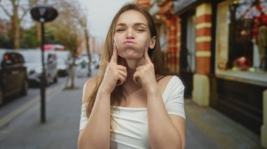 Beyaz omuzlu beyaz elbiseli genç kadın, şehrin caddesinde tuğla dükkanının önünde yanakları çimdikliyor ve somurtuyor..