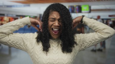 Beyaz kazaklı kadın, bulanık gezginlerin ortasında yoğun havaalanı terminalinde kulaklarını kapatıyor..