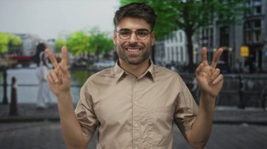 Şehir caddesindeki kanalda barış işareti gösteren bir adam su boyunca yürüme mesafesinde arkasında bulanık binalar, umut..