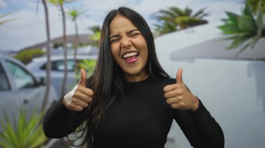 Parlak gökyüzünün altında, arabalarla ve yeşilliklerle çevrili tropikal bir sokak ortamında baş parmağıyla kutlama yapan bir kadın..