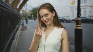 Beyaz elbiseli gülümseyen kadın, kentin işlek kaldırımında el hareketleriyle işlenmiş demir çitin yanında işaret ediyor; davet.