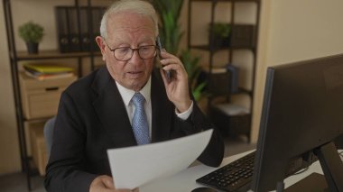 Takım elbiseli üst düzey bir adam ofis masasında bilgisayarlı ve kitaplıklı belgeleri incelerken iş görüşmesine katılmış..