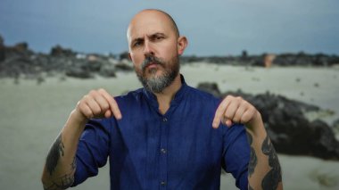 Sakallı bir adam bulutlu bir gökyüzünün altında mavi bir gömlek giyerken kayalık bir sahilde güven içinde el kol hareketi yaparak engebeli bir çevreyi gösteriyor..