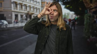Uzun saçlı sarışın adam, kalabalık bir şehir caddesinde dikilirken el üstünde işaret dili gösteriyor..