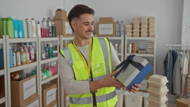 Yansıtıcı yelek giyen genç bir İspanyol, bağış merkezinde bir hediye kutusu tutuyor. Etrafında bir sürü malzeme var..