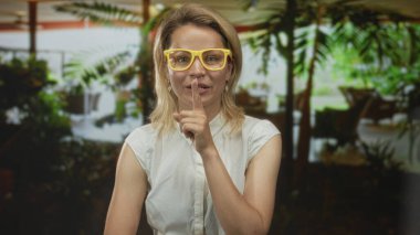 Sarı gözlüklü sarışın kadın, parmağını onun tarafına doğru uzatıyor ve otel binasında gülümsüyor; davetiye..