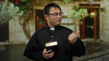 Papaz kıyafeti giyen, elinde kitap tutan, arka planda bulanık kırsal binayla dışarıda duran genç adam.