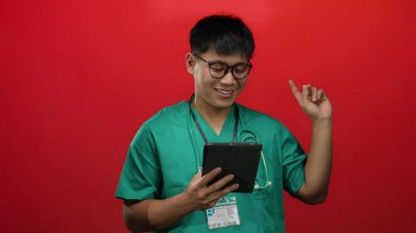 Gülümseyen adam yeşil önlük giyip kırmızı zemine tablet tutarak olumlu tıbbi bilgi ya da başarı öneriyor.