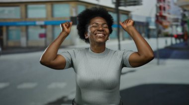 Arka planda bulanık binalar olan bir şehir caddesinde neşeli bir şekilde gülümseyen genç Afro-Amerikalı kadın gündüz vakti kollarını neşeyle kaldırıyor..