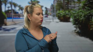 Sarışın bir kadın mavi kapüşonlu ceketini güneş ışığında kaldırımda palmiye ağaçlarıyla birlikte kenara çekiyor..