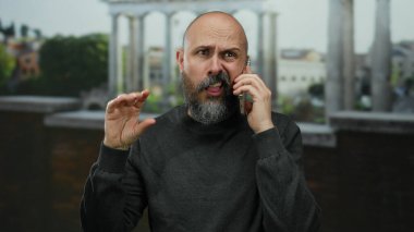 Cep telefonuyla Roma harabelerine karşı konuşan sakallı kel bir adam, endişeli görünüyor ve tarihi mekanda el kol hareketi yapıyor..