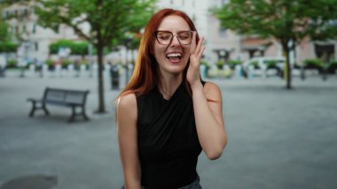 Ağaçlarla dolu bir caddede gülümseyen bir kadın, kızıl saçlı ve gözlüklü, şehir ortamında güzelliği ve zarafeti somutlaştıran bir kadın..