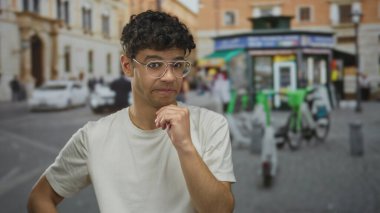 Elleri çenesinde gözlüklü bir adam caddede duruyor. Bulanık dükkanları ve park etmiş bisikletleri var. Belirsizlik düşüncesi..