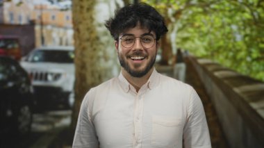 Güneşli bir sokakta gülümseyen sakallı ve gözlüklü genç bir adam. Arka planda ağaçlar ve arabalar var..