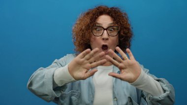 İspanyol, orta yaşlı, kıvırcık saçlı bir kadın kameraya, izole edilmiş mavi bir arkaplana, gözlüklü ve pozitif enerji gösteren günlük gömlekli bir kadına coşkuyla el sallıyor..