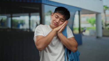 Şehir sokaklarında genç bir adam, beyaz tişört ve mavi sırt çantasıyla ellerine karşı uyku jesti yapıyor. Gün içinde sakin bir ifadeyle gülümsüyor.