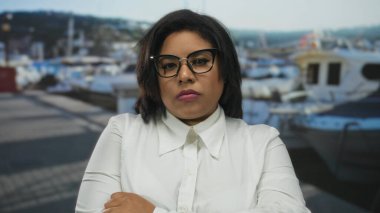 Gözlüklü, beyaz tişörtlü, teknelerin yanında duran bir kadın gündüz vakti bir limanda yakalanmış. Arkaplanı bulanık, kendine güveni tam bir ifade sergiliyor..