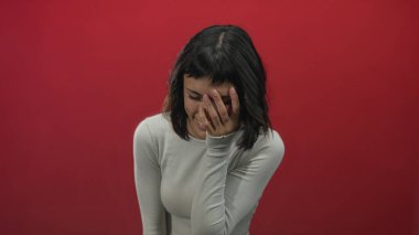 Uzun kollu bluzlu genç İspanyol kadın elleriyle ağzını kapatır ve kırmızı stüdyoda başını eğer; mutluluk..