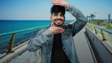 Kotlu bir adam güneşli bir günde deniz kenarında neşe içinde poz veriyor..