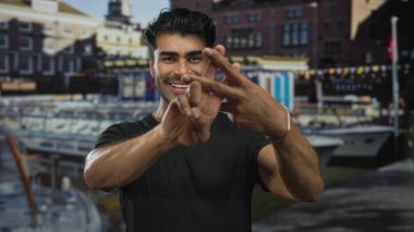 İspanyol asıllı genç adam hareketli bir limanda limanda limanlı bir yatın yanında hashtag jesti yapıyor; sosyal medya bağlantısı mutluluğu.