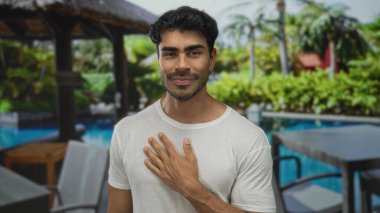 Beyaz tişörtlü adam oteldeki yemyeşil tropikal havuz manzaralı sazlı bir çardağın altındaki havuz kenarındaki sandalyeyi işaret ediyor..