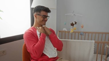 Yatak odasında kucağında bir bebek elbisesi tutan genç bir adam, sıcak aydınlatılmış bir çocuk yuvasında ebeveynlik için mutluluk ve beklenti gösteriyor..