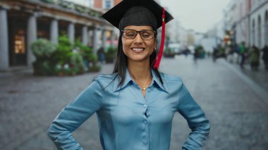 Şehir ortamında gülümseyen akademik şapkalı bir kadın mezuniyeti sembolize ediyor, eğitimsel başarıyı sokakta kendinden emin bir şekilde kutluyor genç Latin profesyonel