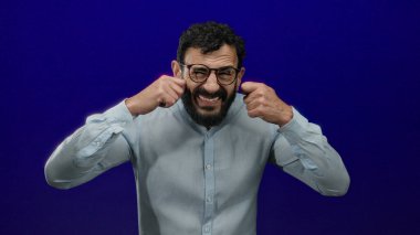 Sakallı, duygusal görünen, gözlüklü ve açık renk gömlekli bir adam mavi arka planda gözlerinin yanındaki elleri kullanarak üzüntü ve hayal kırıklığını çağrıştırıyor..