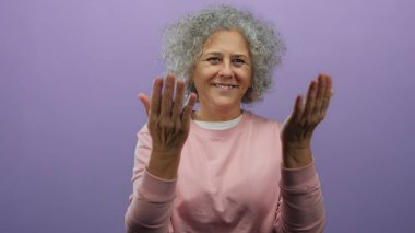 Gri saçlı yaşlı bir kadın, pembe bir süveter giyen ve nazikçe gülümseyen, izole bir mor duvar arka planına el hareketi yapıyor..