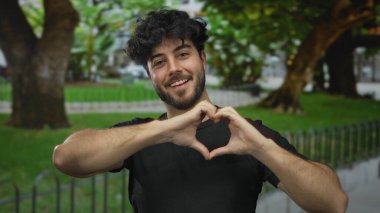 Sakallı genç adam, açık yeşil bir parkta gülümserken kalp hareketi yapıyor, sevgi ve mutluluk taşıyor..