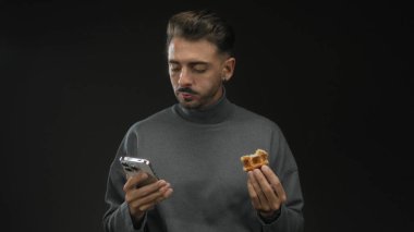 Siyah stüdyo ortamında akıllı telefon tutarken waffle yiyen adam; çoklu görev kolaylığı rahatlaması.